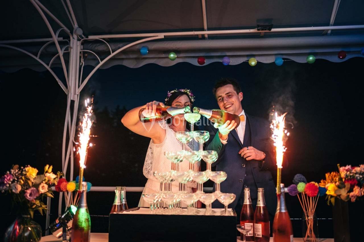 Un couple remplit une pyramide de coupes avec du champagne, entouré de décorations festives et de feux d'artifice.