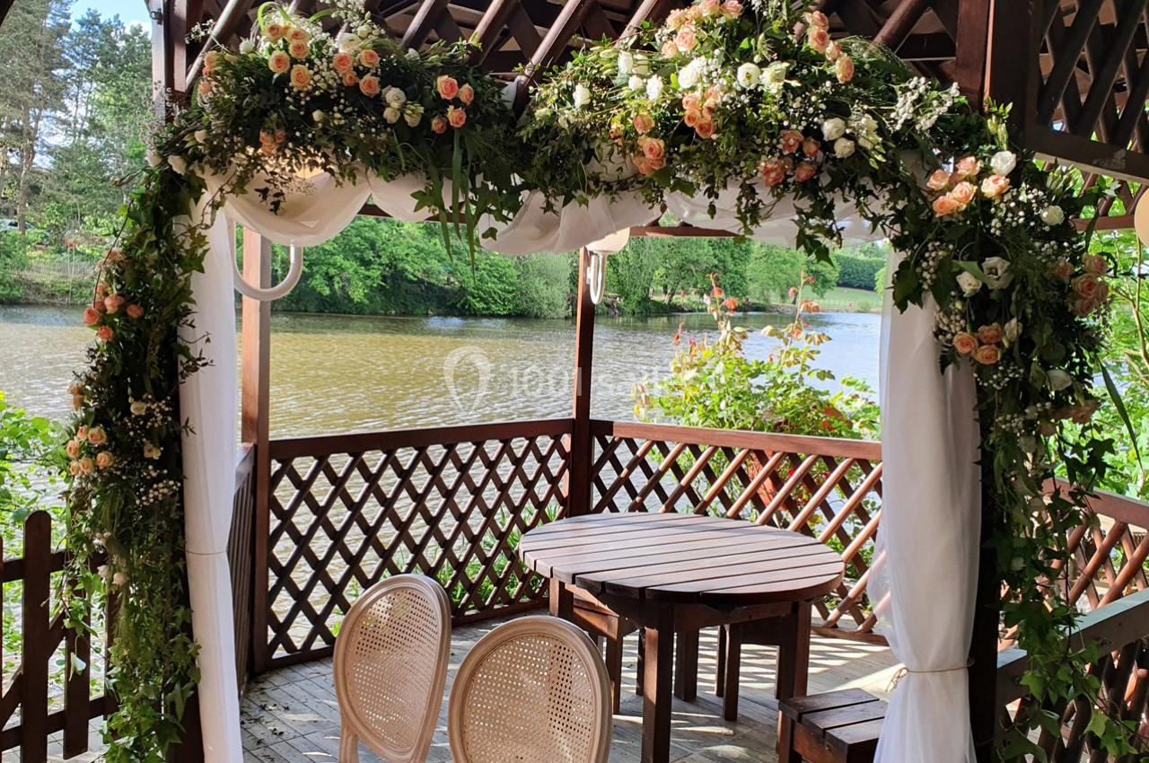 Pergola en bois décorée de fleurs et de voilages blancs, avec table et chaises, au bord d'un étang entouré de verdure.