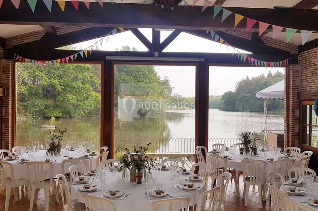 Salle de réception décorée avec des guirlandes colorées, tables dressées, vue sur un lac et des arbres à travers de grandes…