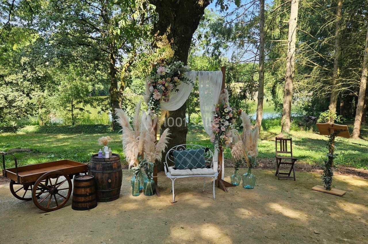 Décoration champêtre avec arche florale, banc blanc, tonneaux et chariot en bois, dans un cadre boisé ensoleillé.