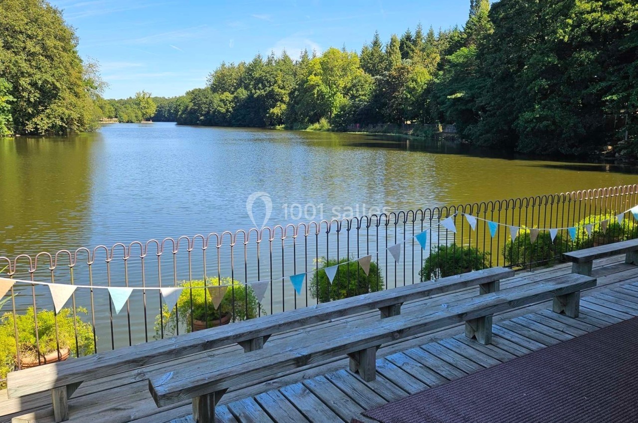 Vue d'un lac entouré d'arbres avec un ponton en bois, des bancs et des guirlandes colorées accrochées à une barrière.