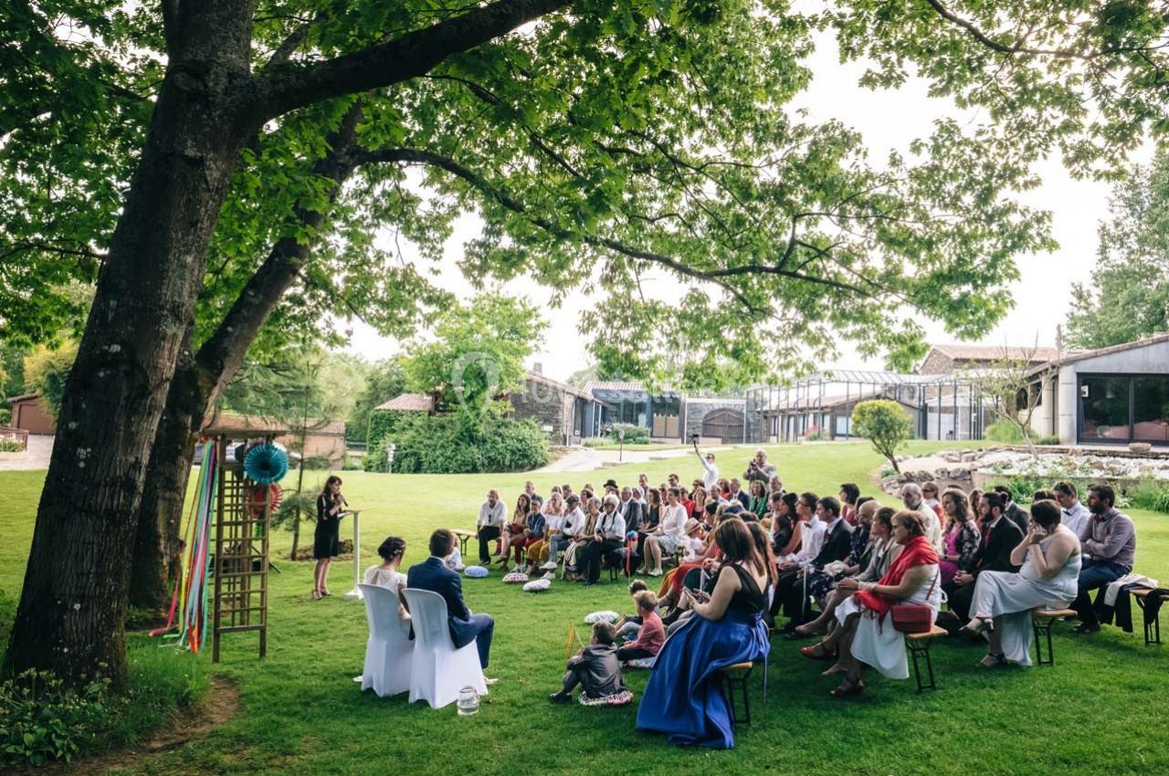Un groupe de personnes assises en plein air sous un grand arbre, écoutant une femme debout qui s'exprime.