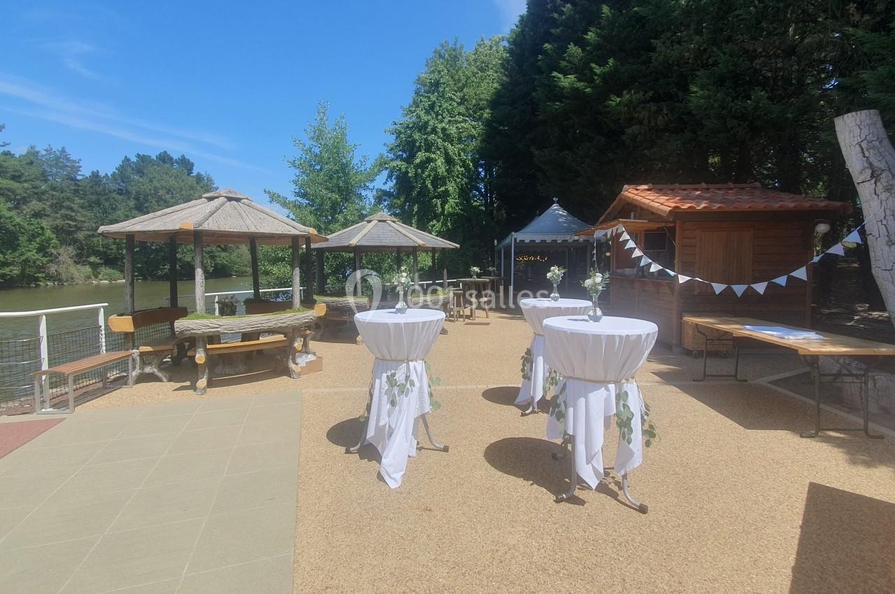 Espace extérieur aménagé avec tables hautes décorées, parasols et cabanes en bois près d'un plan d'eau.