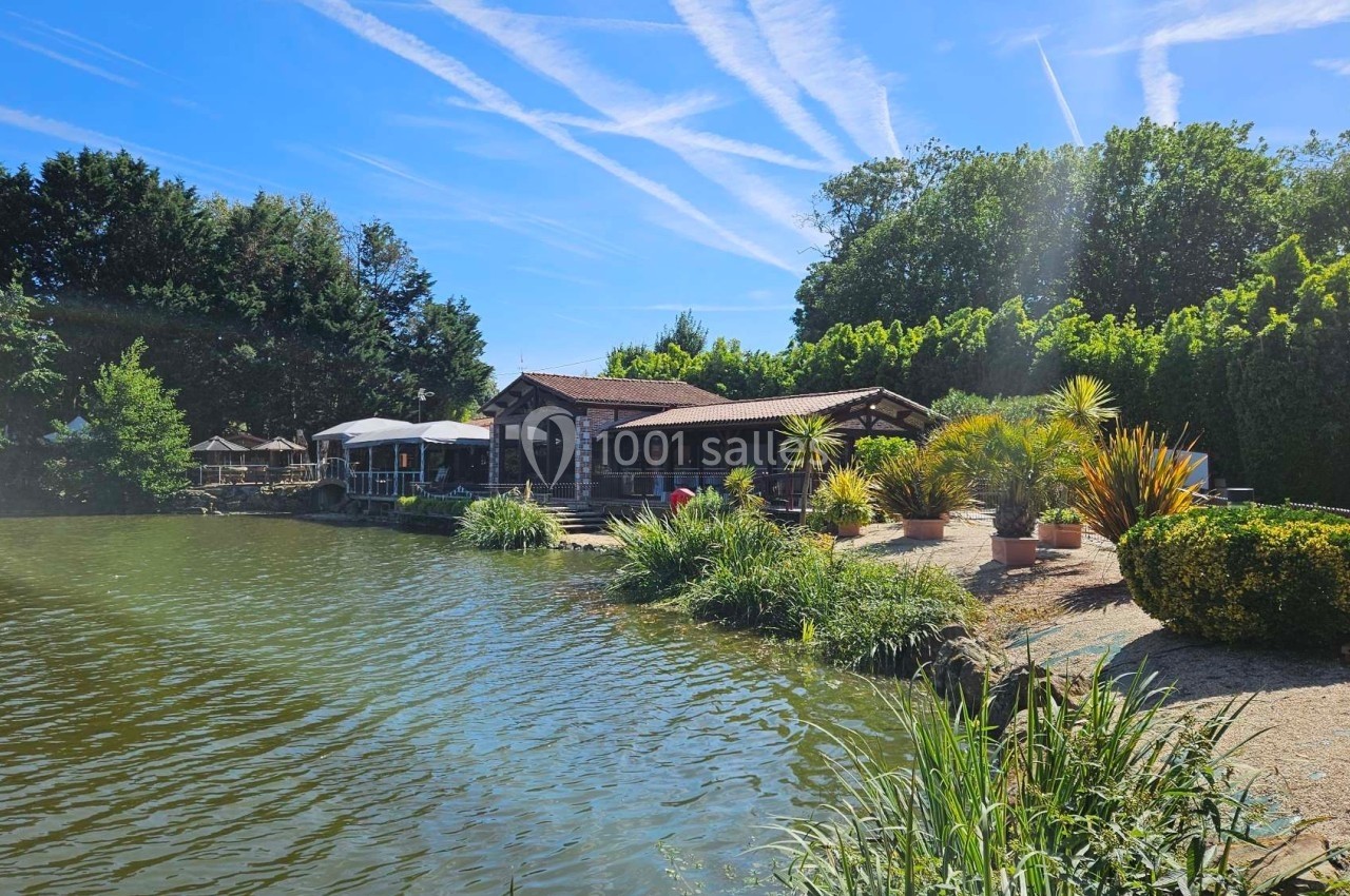 Bâtiment en bois avec terrasse au bord d'un étang, entouré de végétation et éclairé par un ciel ensoleillé.