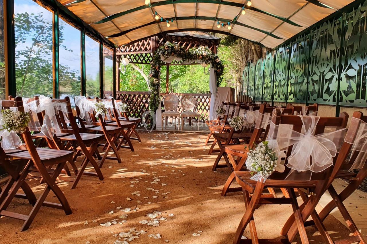 Chaises en bois décorées de rubans et de fleurs blanches disposées dans une allée menant à une arche florale dans un espace…