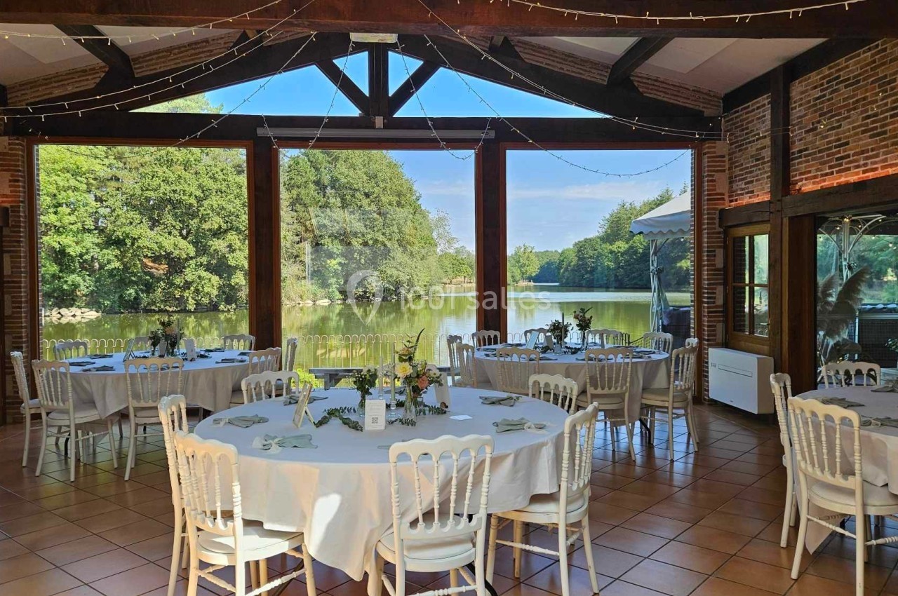Salle de réception lumineuse avec vue sur un lac, tables rondes dressées et décorations florales.