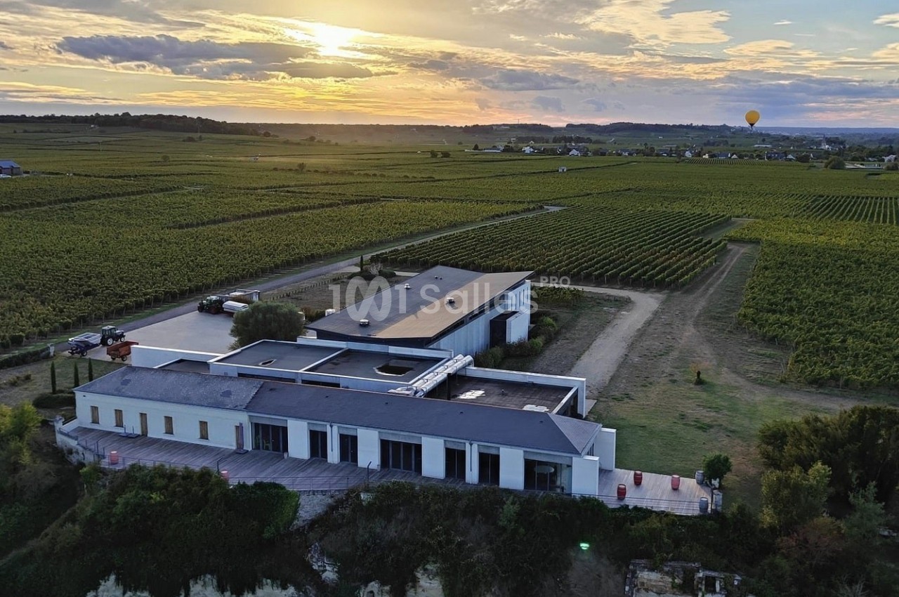 Bâtiment moderne entouré de vignobles au coucher du soleil, avec une montgolfière visible à l'horizon.