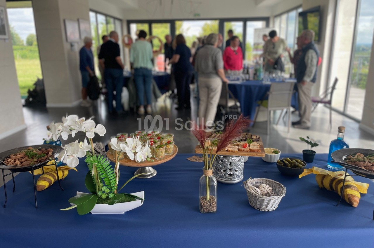Table garnie de plats et décorations florales au premier plan, avec des personnes discutant en arrière-plan dans une salle…