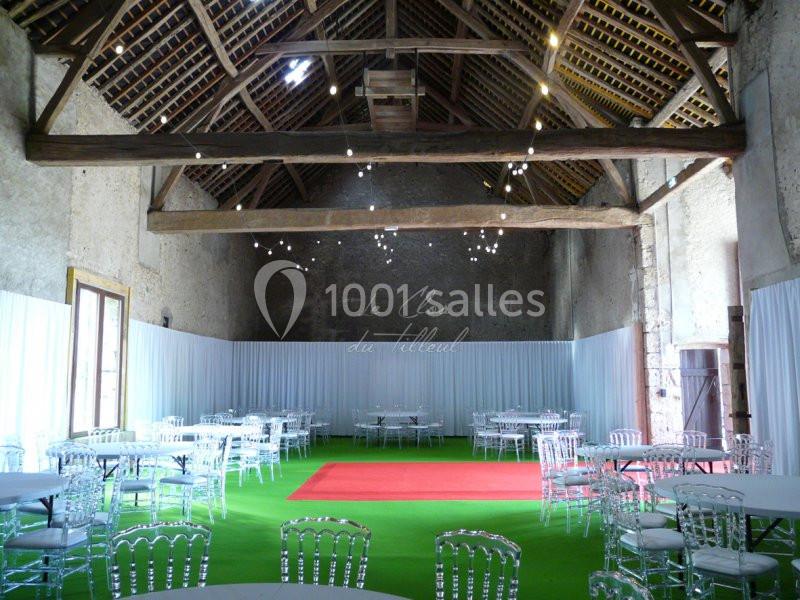 Salle de réception avec charpente en bois apparente, tables rondes et chaises transparentes sur un sol vert.