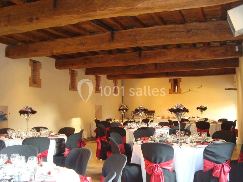 Salle de réception avec tables rondes dressées, nappes blanches, chaises noires ornées de nœuds rouges et poutres apparentes.