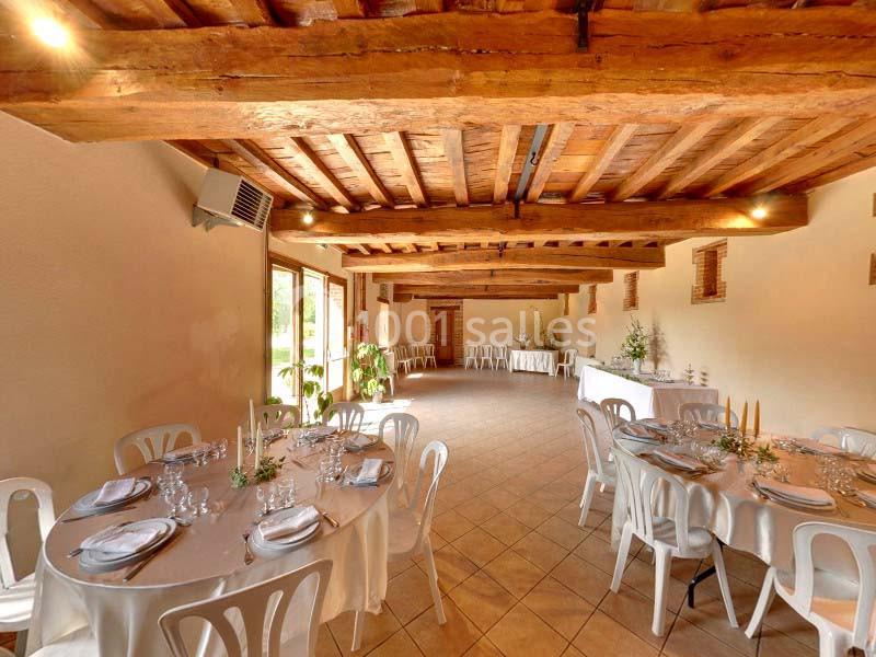 Salle de réception avec tables rondes dressées, chaises blanches et poutres apparentes en bois.