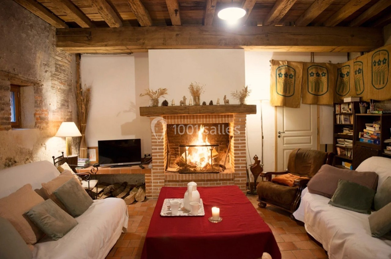 Salon chaleureux avec cheminée allumée, poutres apparentes, canapé, fauteuils et table basse décorée.