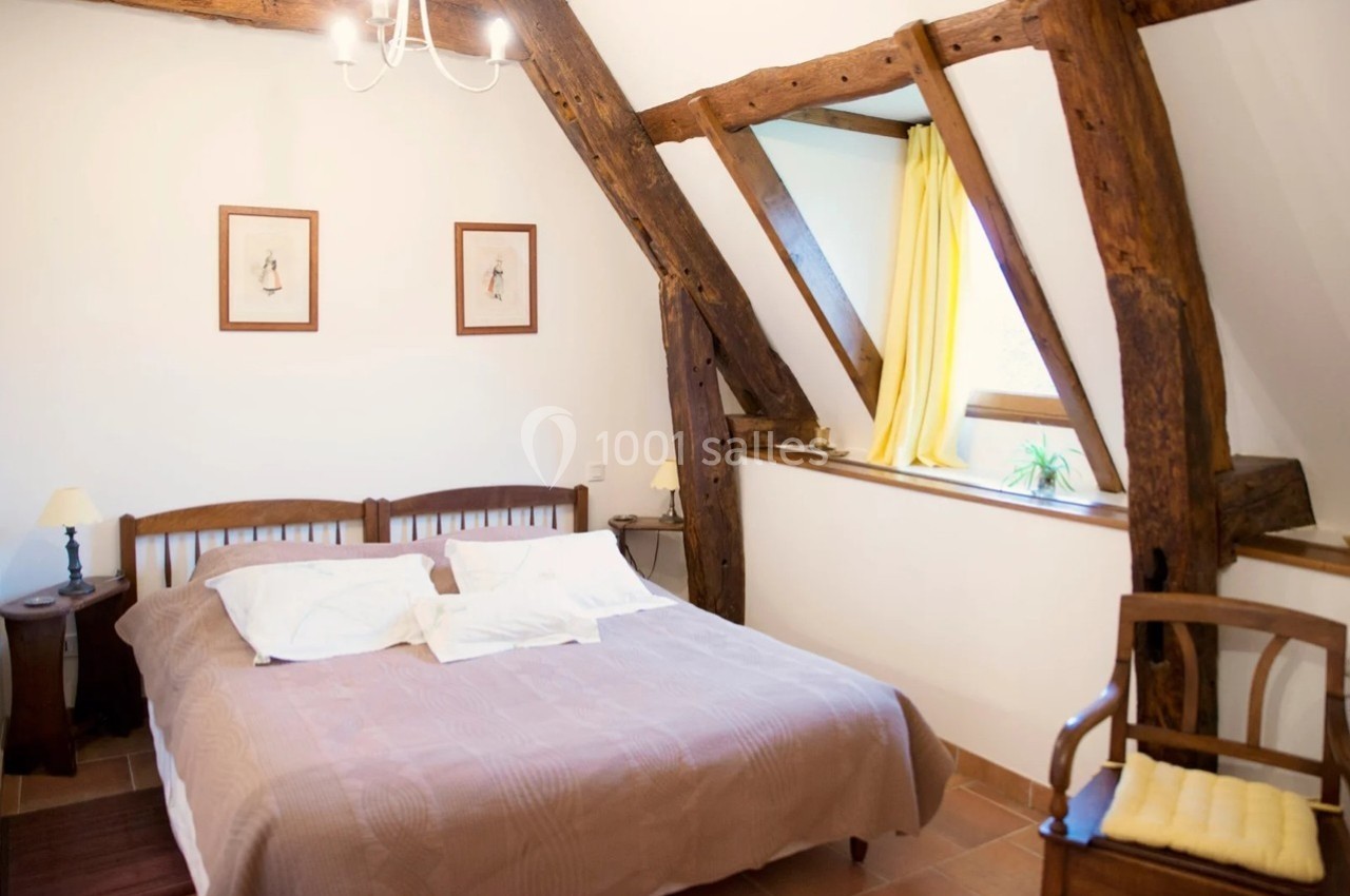 Chambre lumineuse avec poutres apparentes, lit double, fenêtres avec rideaux jaunes et décoration sobre.