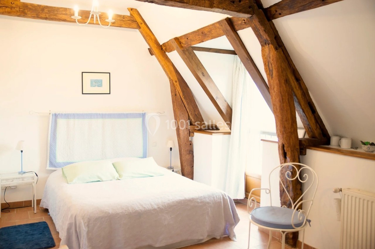 Chambre lumineuse avec poutres apparentes, lit double, chaise en métal et fenêtre laissant entrer la lumière naturelle.