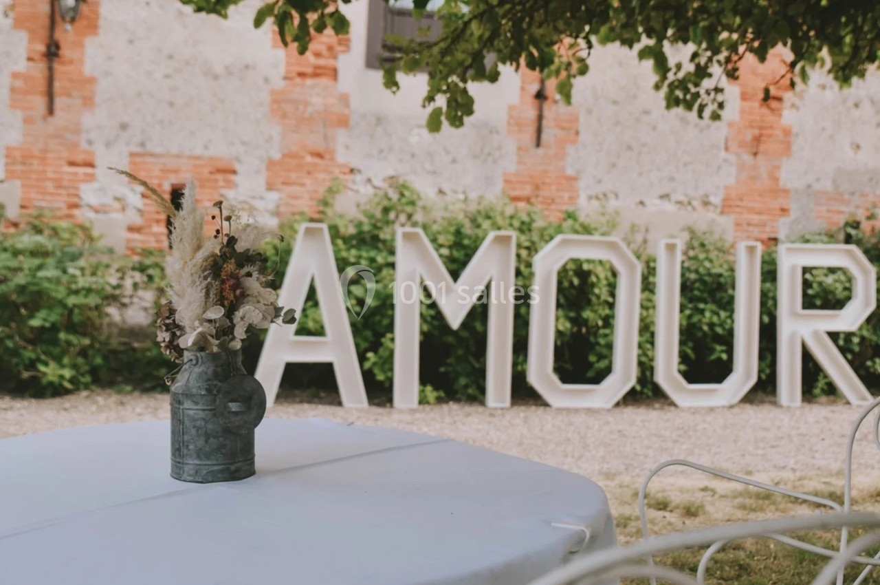 Vase de fleurs séchées sur une table blanche en extérieur, devant des lettres géantes formant le mot ’AMOUR’.