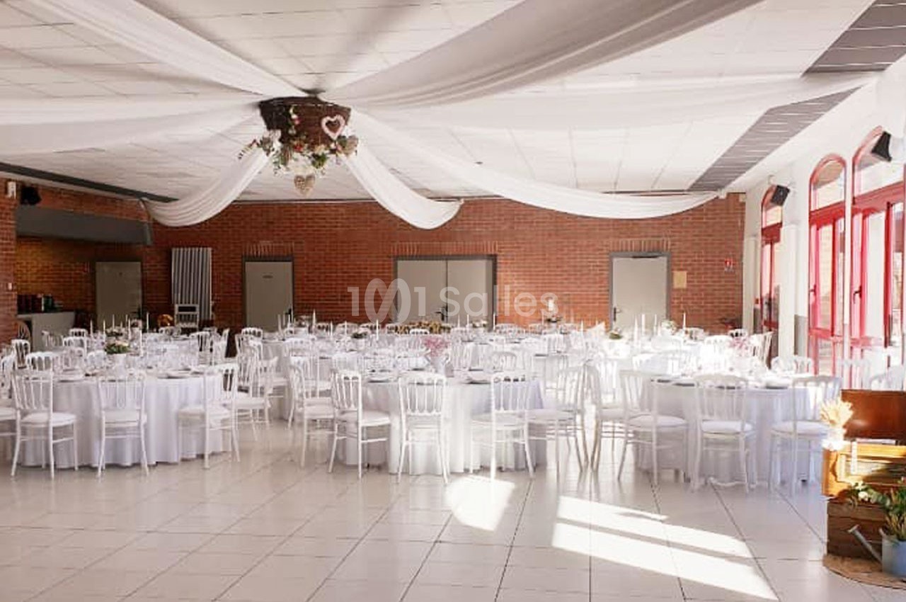 Salle de réception décorée avec des tables rondes dressées, chaises blanches et drapés suspendus au plafond.