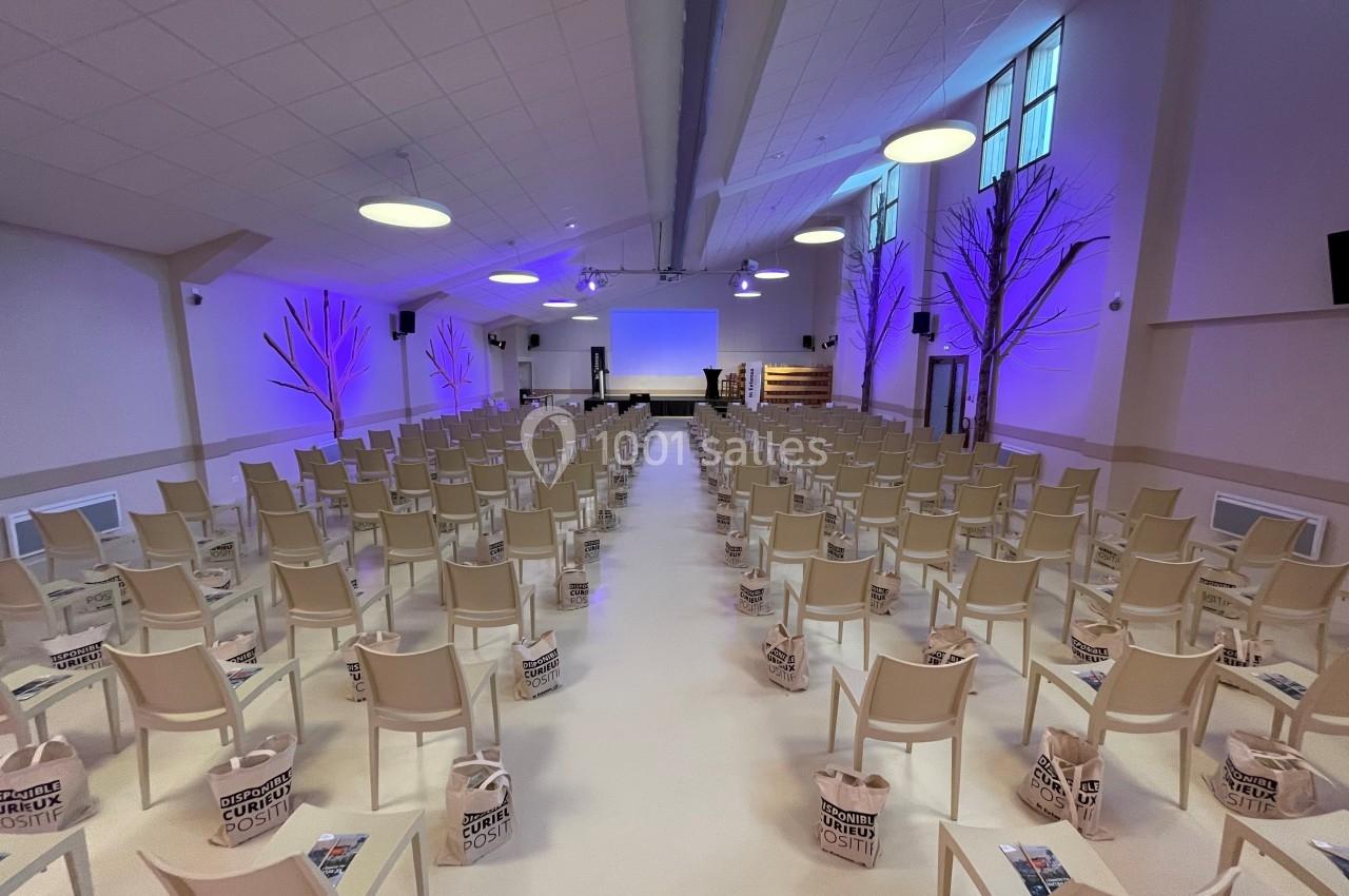 Salle de conférence avec des rangées de chaises alignées, sacs posés au sol et éclairage violet sur les murs.