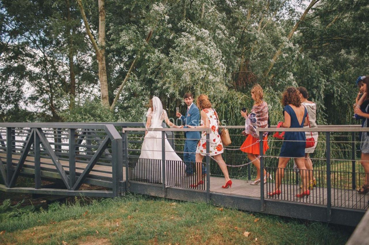 Un couple de mariés traverse un pont en bois, suivi de plusieurs invités vêtus de tenues colorées.