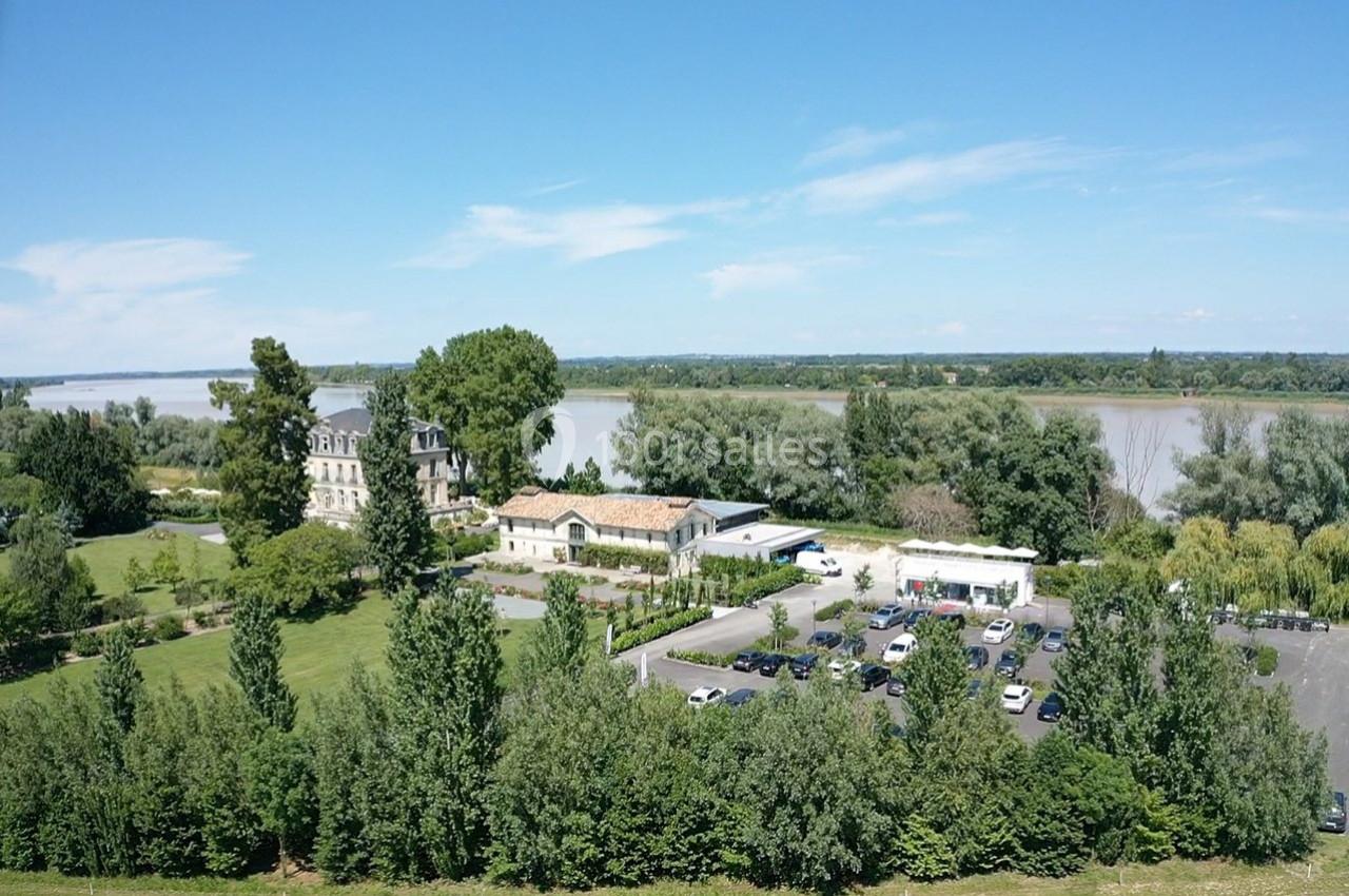 Vue aérienne d'un domaine avec un bâtiment principal, un parking et un jardin, près d'une rivière bordée d'arbres.