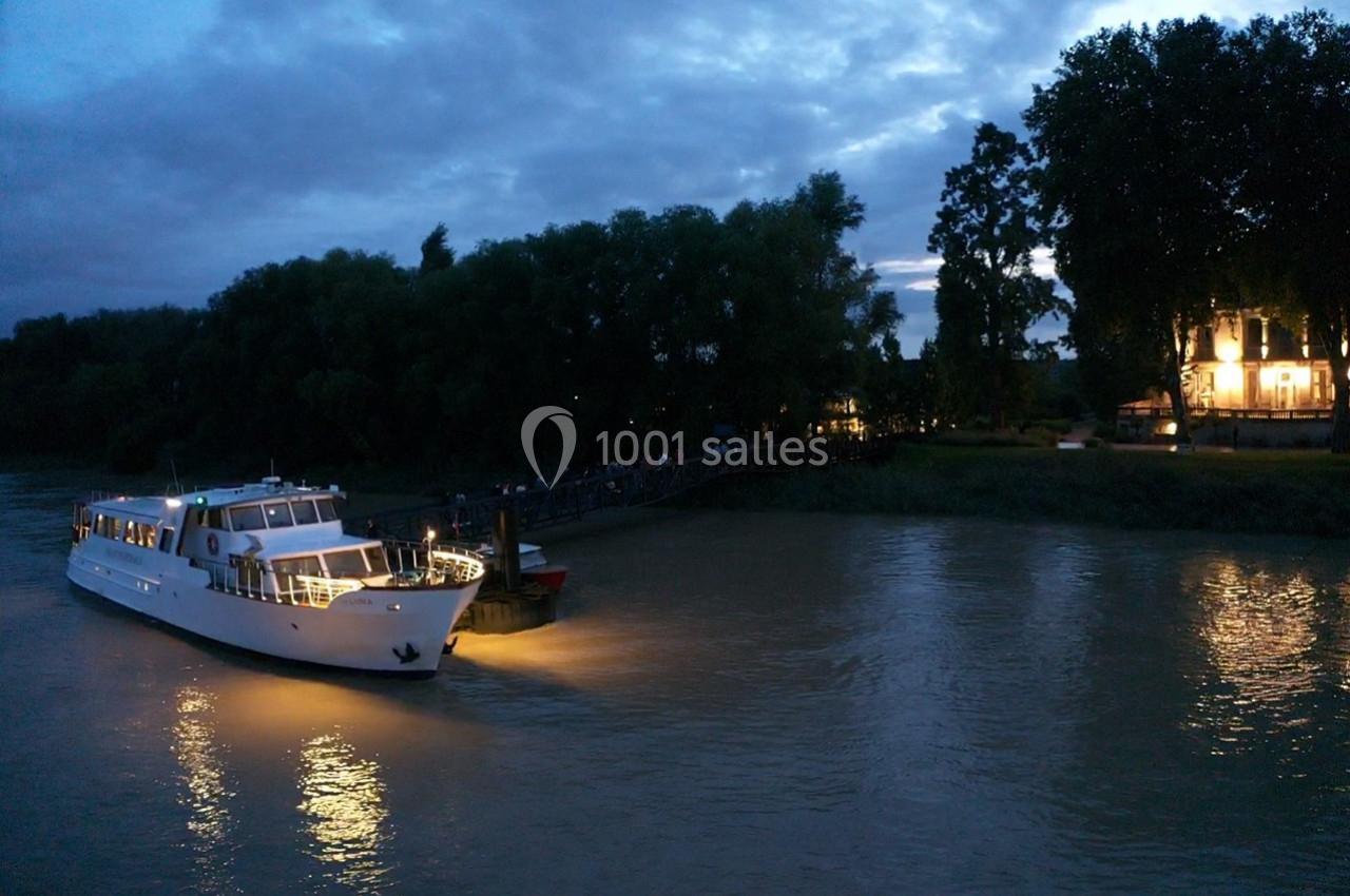 Un bateau éclairé navigue sur une rivière au crépuscule, avec des arbres et un bâtiment illuminé en arrière-plan.