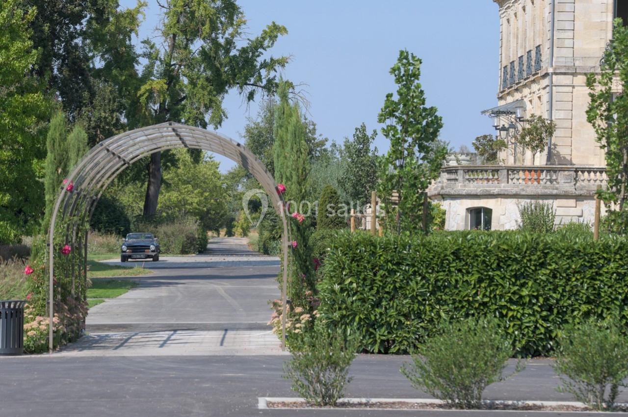 Allée bordée de verdure avec une arche fleurie, menant à un bâtiment en pierre et une voiture au loin.