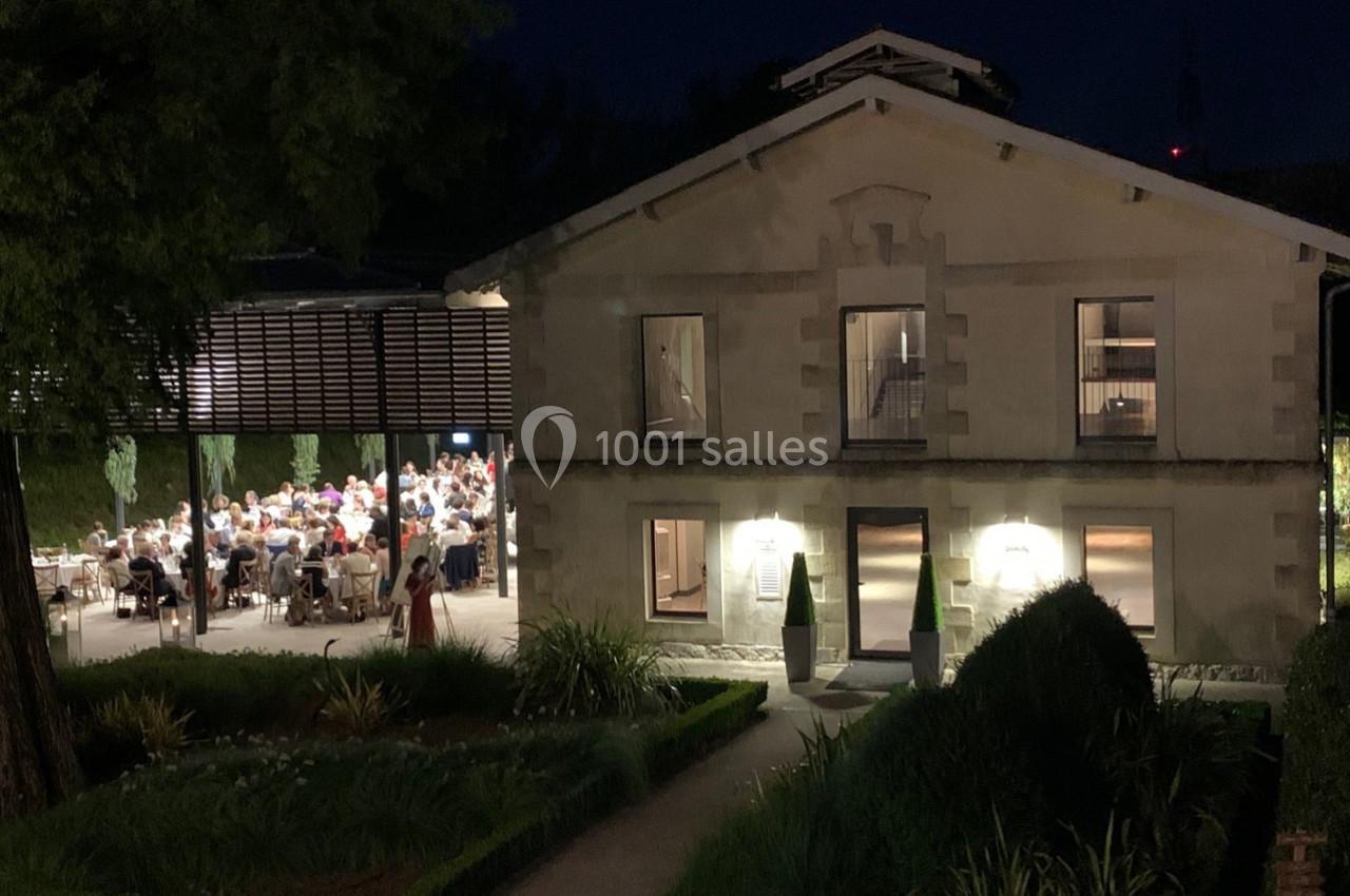 Façade éclairée d'un bâtiment en soirée avec des convives attablés sous une terrasse couverte à côté.
