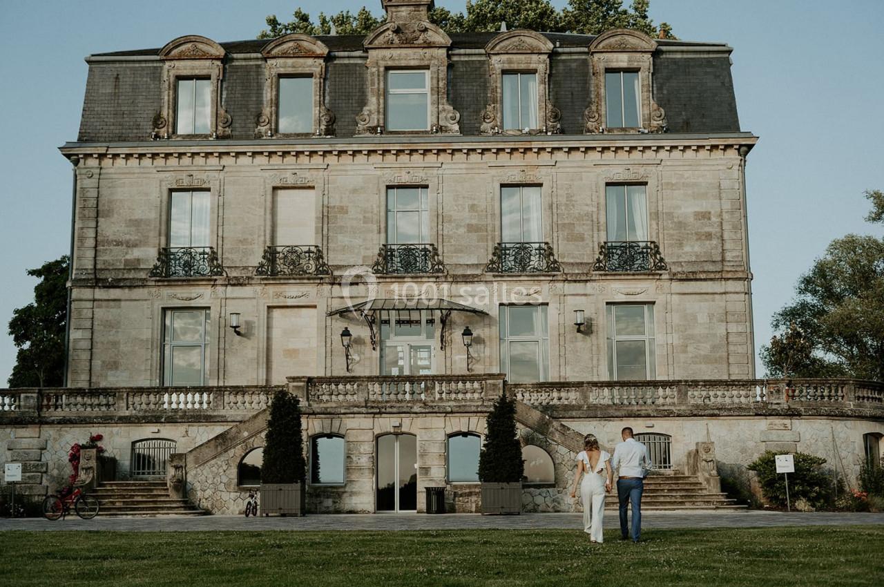 Façade d'un manoir en pierre avec balcons en fer forgé, un couple marchant sur la pelouse au premier plan.