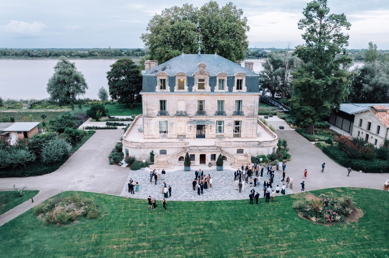 Vue aérienne d'un manoir entouré de jardins, avec des personnes rassemblées sur une terrasse pavée devant.