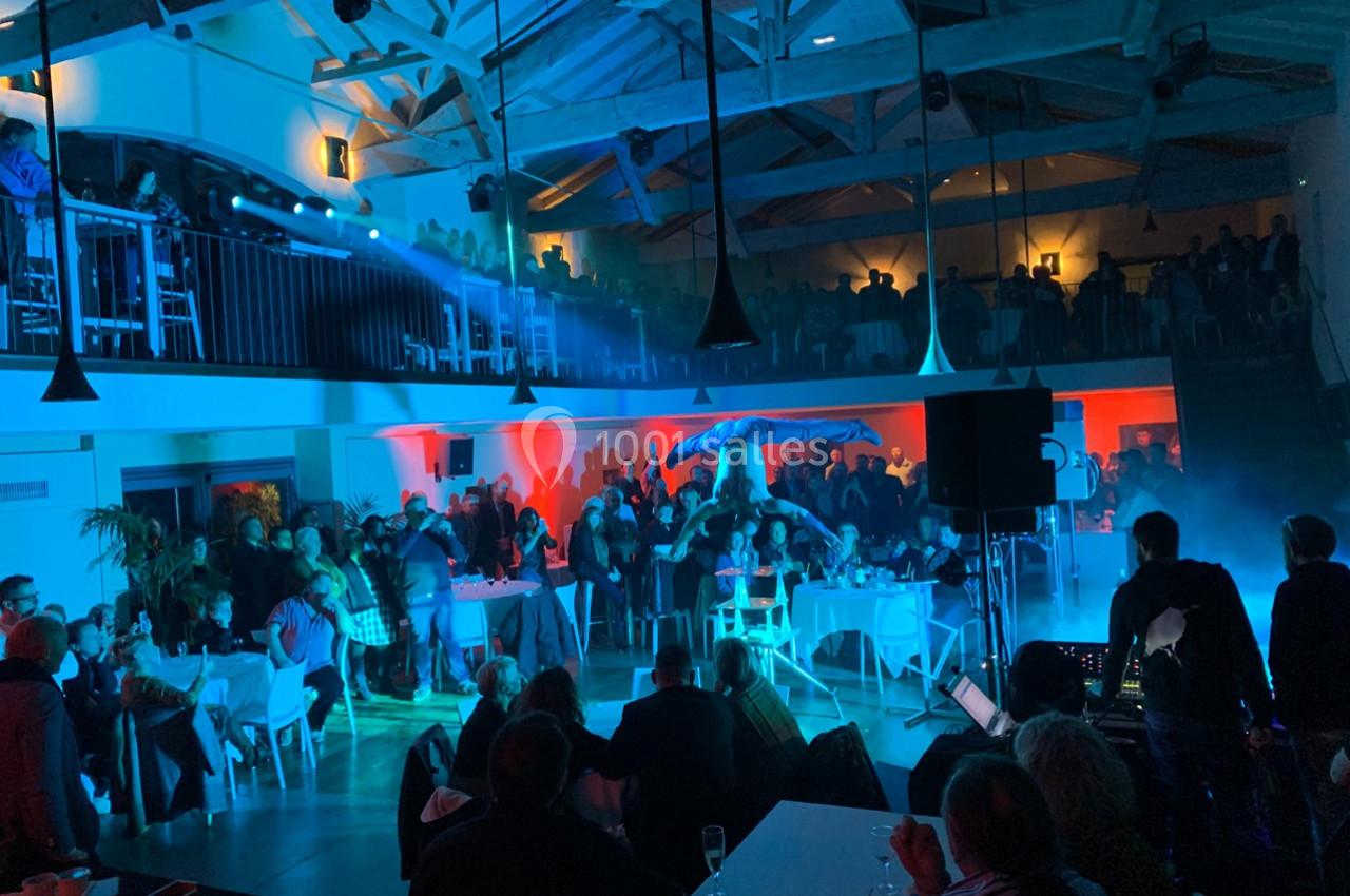 Salle animée avec des invités assis et debout, éclairage coloré et spectacle sur scène sous un parasol.