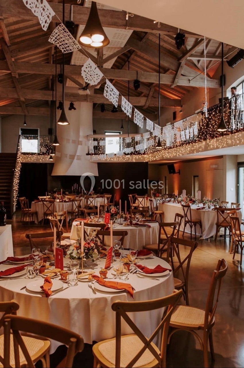 Salle de réception décorée avec des guirlandes lumineuses, des tables rondes dressées et un plafond en bois apparent.