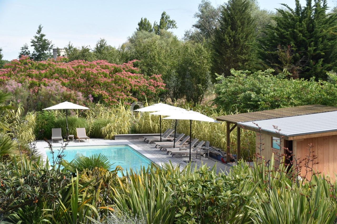 Piscine extérieure entourée de végétation luxuriante, avec chaises longues, parasols et un abri en bois.