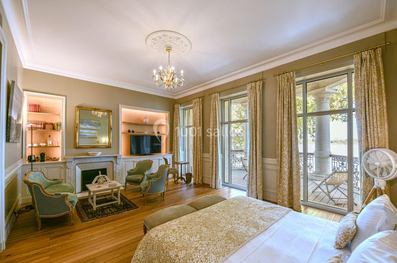 Chambre lumineuse avec lit double, coin salon, cheminée, télévision et grandes fenêtres donnant sur une terrasse.