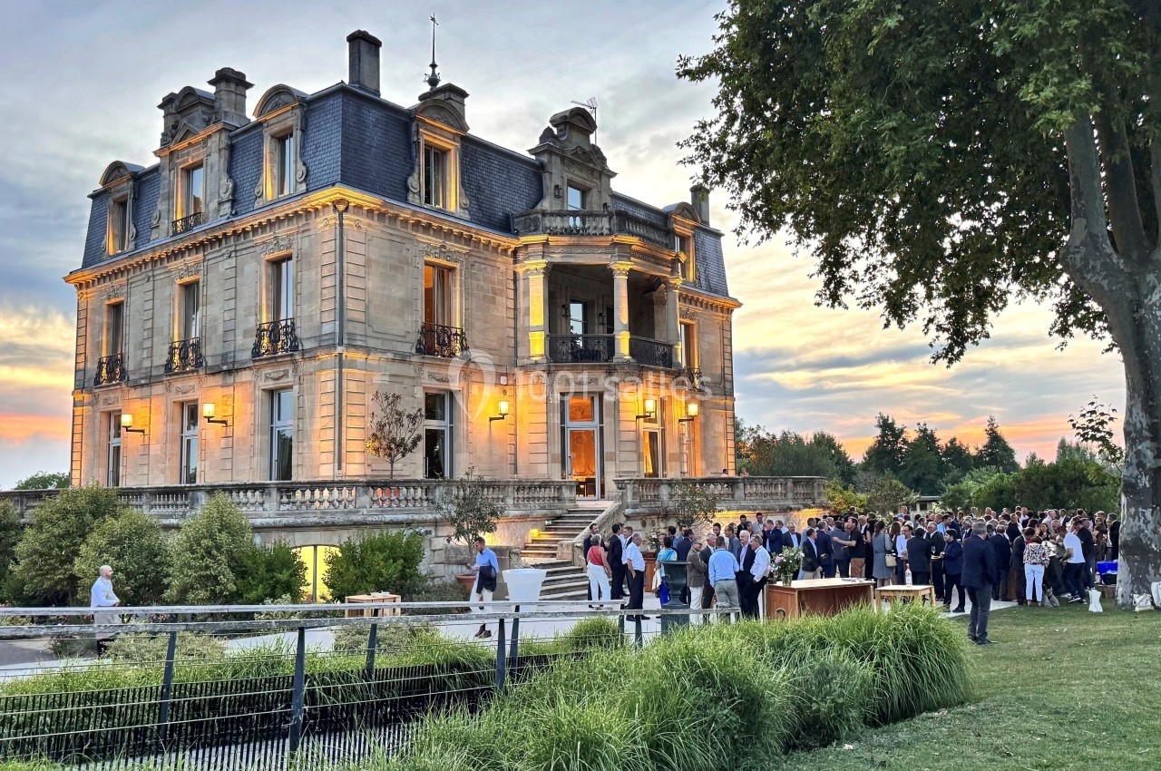 Groupe de personnes rassemblées devant un château éclairé au crépuscule, entouré de verdure.