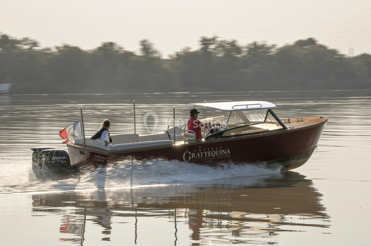 Un bateau à moteur navigue sur une rivière calme, avec deux personnes à bord et des arbres en arrière-plan.