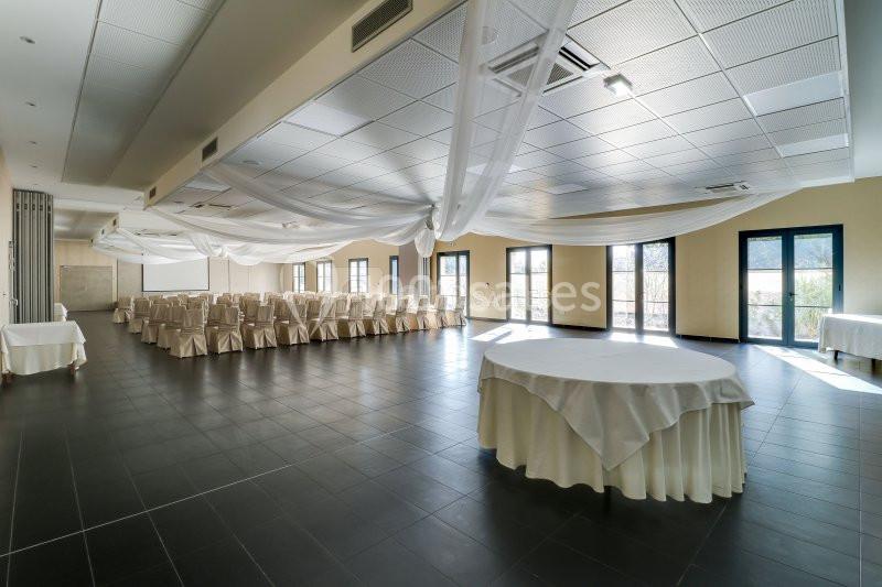 Salle de réception lumineuse avec chaises alignées, tables rondes dressées et grandes fenêtres donnant sur l'extérieur.
