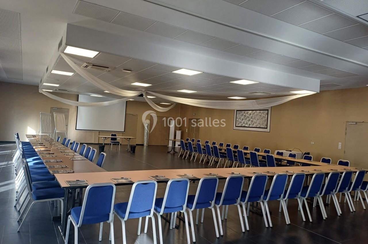 Salle de réunion spacieuse avec des tables disposées en U, des chaises bleues et un écran de projection.
