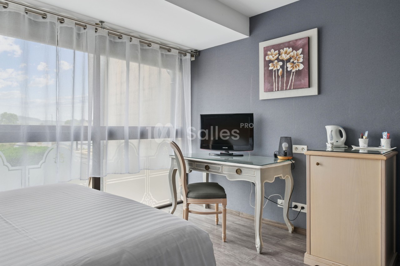 Chambre lumineuse avec bureau, télévision, bouilloire et grande fenêtre avec rideaux translucides.