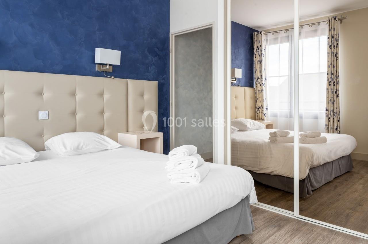 Chambre lumineuse avec un lit double, des draps blancs, des serviettes pliées et un grand miroir reflétant la fenêtre.