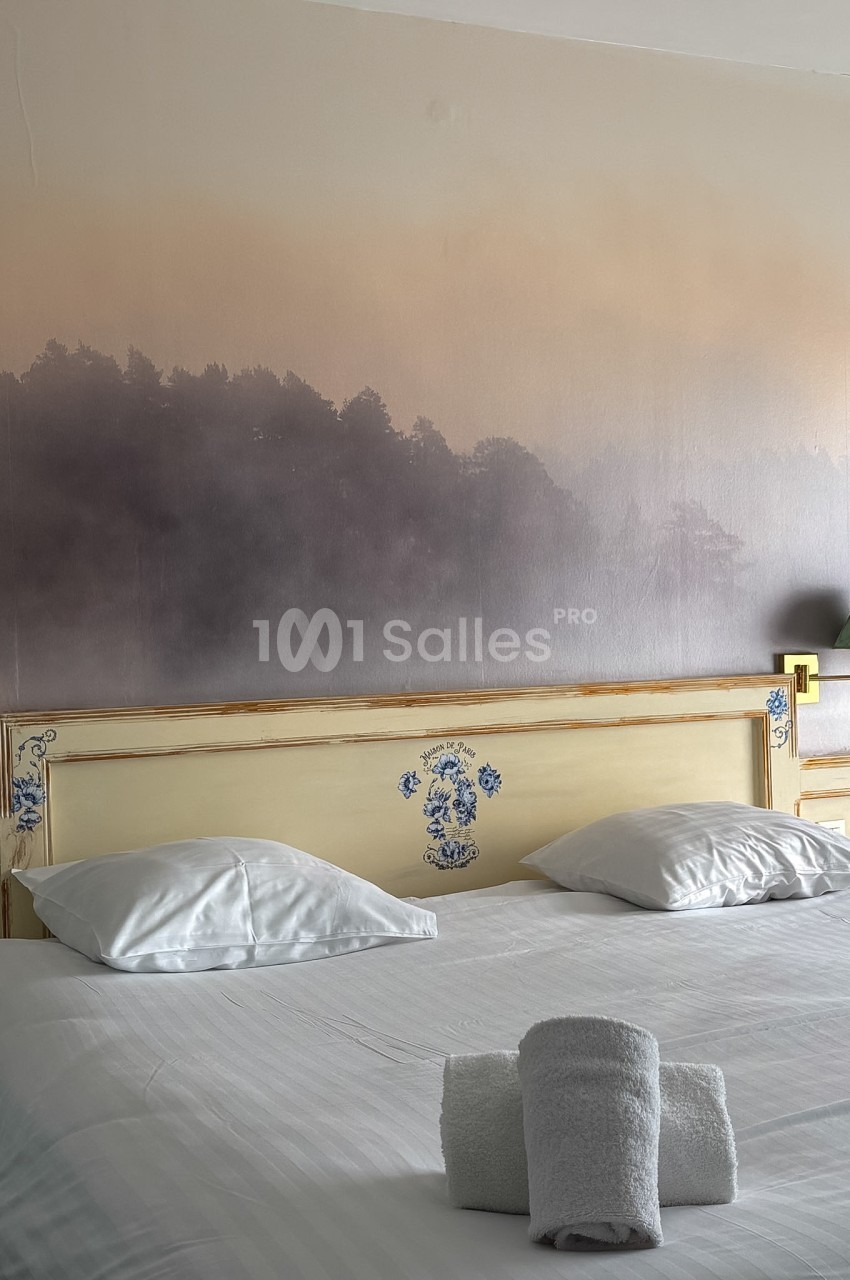 Lit double avec tête de lit décorée, draps blancs et serviettes pliées, devant un mur avec un motif de forêt brumeuse.