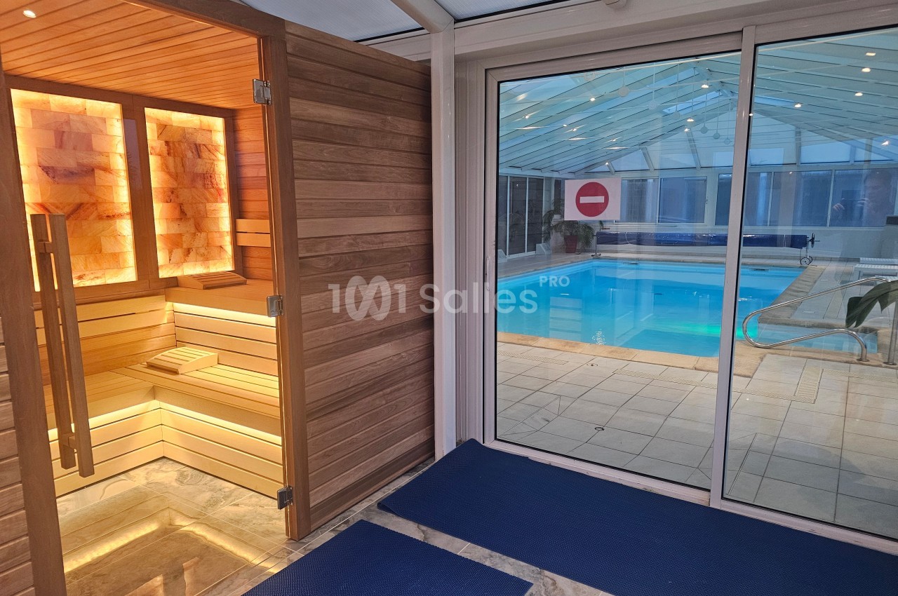 Vue d'un sauna en bois éclairé à gauche et d'une piscine intérieure derrière une baie vitrée à droite.