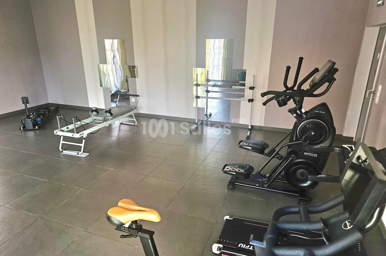 Salle de sport avec tapis de course, vélo d'appartement, rameur et miroirs sur un mur beige.