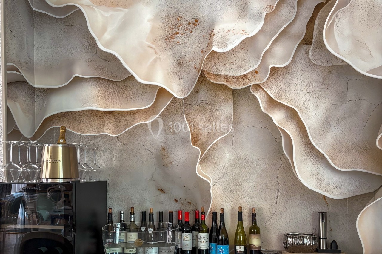 Mur texturé beige évoquant des formes organiques, avec un bar en premier plan contenant bouteilles et verres.