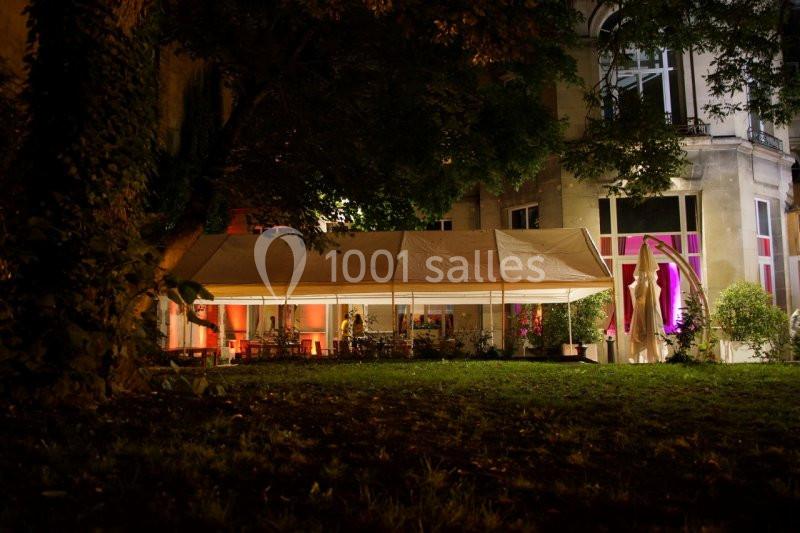 Tente éclairée installée dans un jardin, près d'un bâtiment illuminé, entouré d'arbres en soirée.
