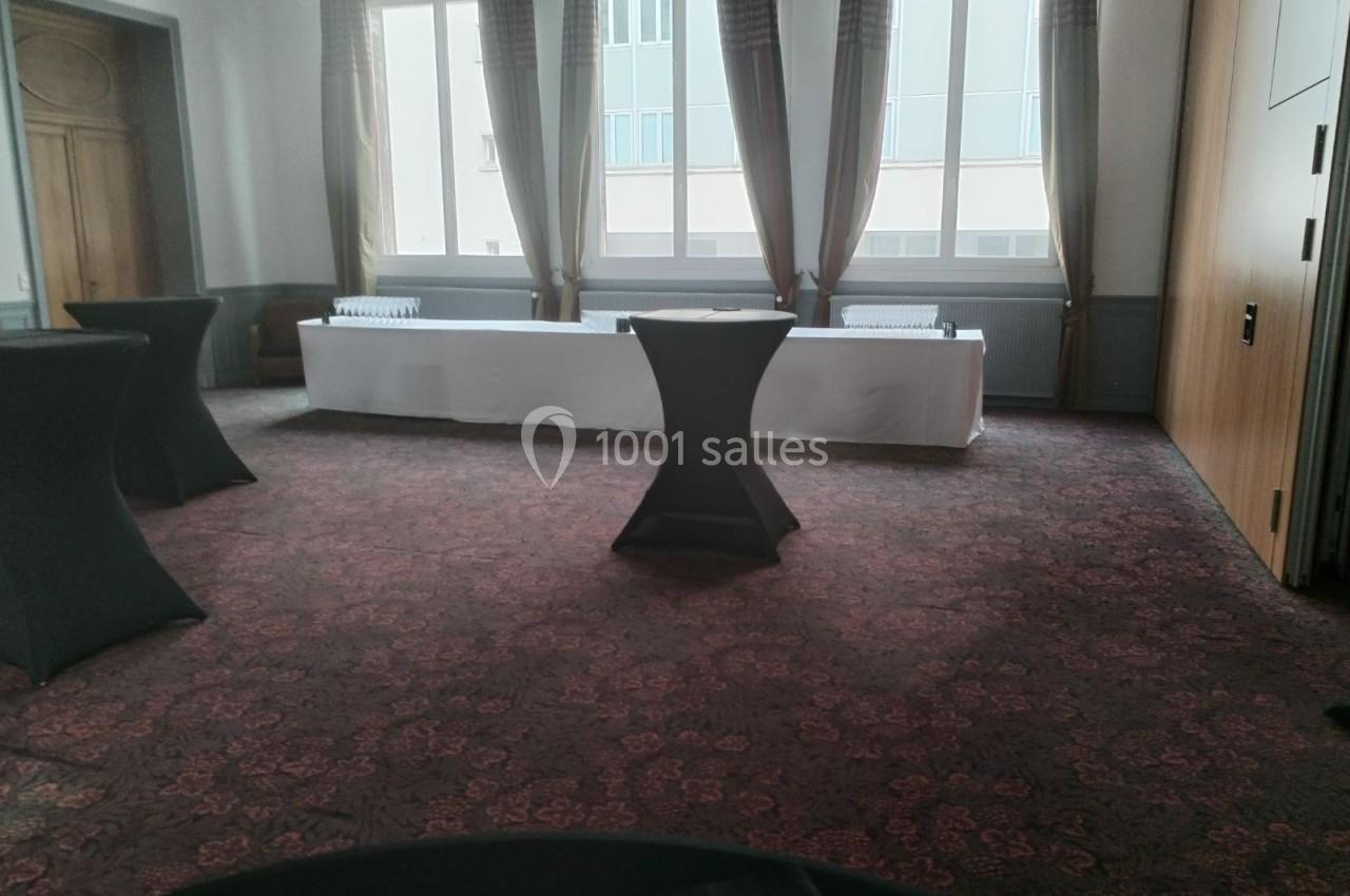 Salle vide avec moquette rouge, tables hautes noires et une longue table blanche près des fenêtres.