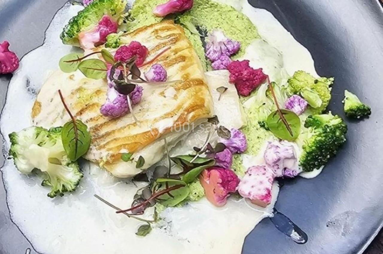 Filet de poisson grillé servi avec une sauce crémeuse, brocolis et choux-fleurs colorés sur une assiette noire.