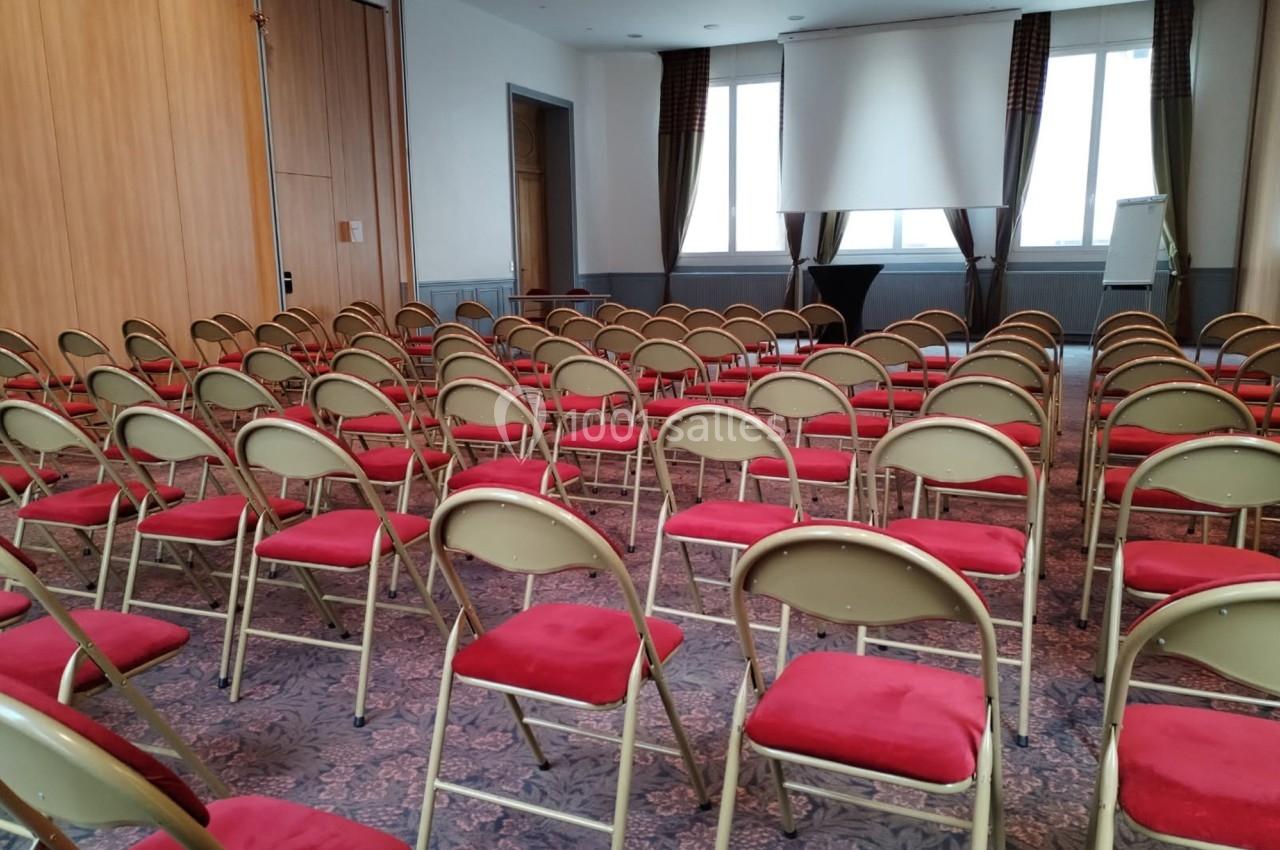 Salle de conférence vide avec des rangées de chaises pliantes rouges, un écran de projection et un chevalet de présentation.