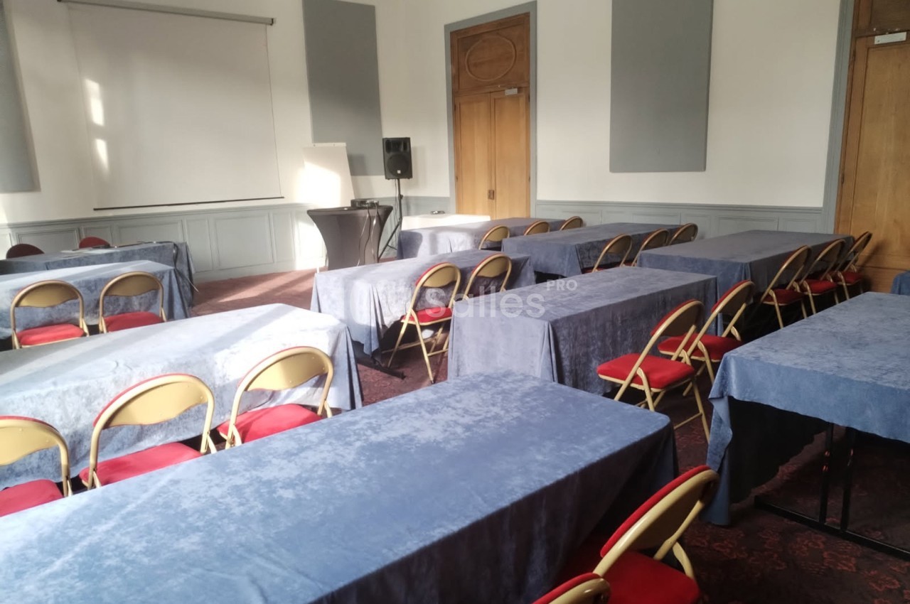Salle de réunion avec des tables recouvertes de nappes bleues, des chaises pliantes et un projecteur près d'un écran blanc.