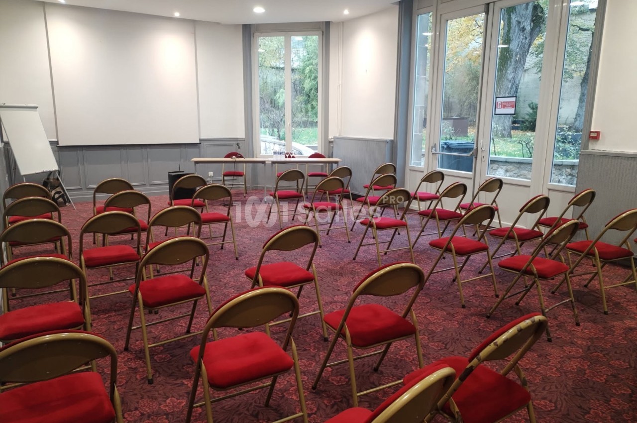 Salle de réunion avec des chaises pliantes rouges disposées en rangées face à une table et un tableau blanc.