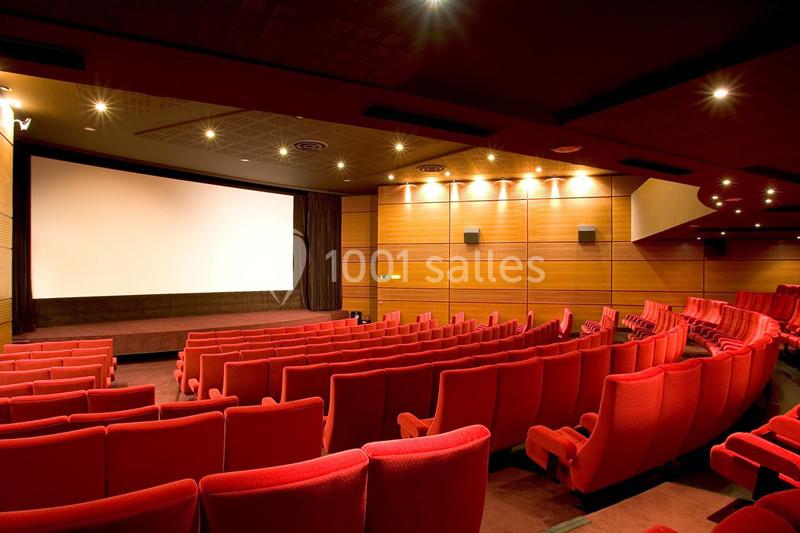 Salle de cinéma vide avec des rangées de sièges rouges, un écran blanc et des murs en bois éclairés.