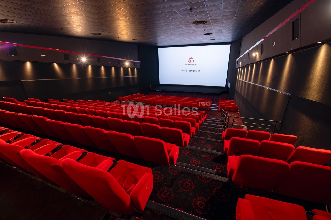 Salle de cinéma vide avec des rangées de sièges rouges et un écran blanc allumé au fond.